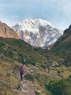 two hikers trekking up a mountain trail towards a snow - capped mountain range