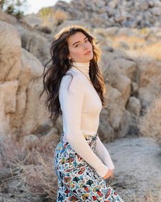 a woman standing in front of rocks wearing a white turtle neck sweater and floral print skirt