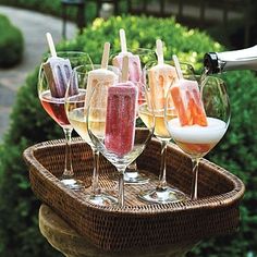 wine glasses filled with different types of drinks sitting on top of a wooden tray in front of bushes