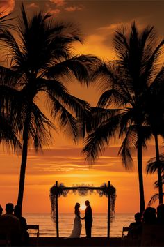 a bride and groom stand under palm trees at sunset on the beach as the sun sets