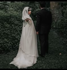 a man and woman dressed in wedding attire walking through the woods together, holding hands