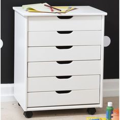 a white dresser with five drawers in front of a black wall and toys on the floor