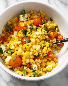 a white bowl filled with corn, tomatoes and basil on top of a marble counter