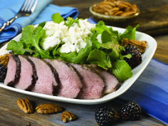 a white plate topped with meat and salad on top of a wooden table next to nuts