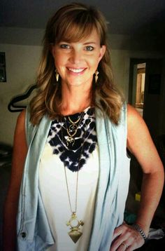 a woman standing in a room wearing a white top and blue vest with gold necklaces