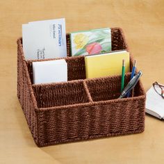 a brown basket with some books and pens in it on a wooden table next to a pair of glasses