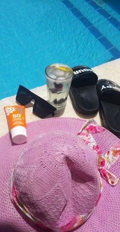 a pink hat sitting on top of a table next to a swimming pool with sun glasses and sunscreen