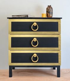 a black and gold chest of drawers with two rings on each drawer, next to a vase