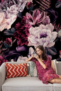 a woman sitting on a couch in front of a floral wallpaper with purple flowers