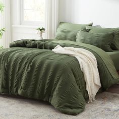 a bed with green comforter and pillows in a room next to a plant on the floor