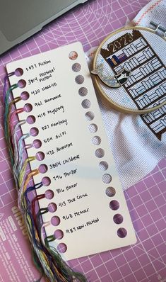 a cross stitch book next to a laptop on a pink table with scissors and thread