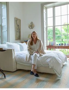 a woman sitting on top of a white bed in a bedroom next to two windows