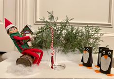 an elf is sitting in a chair next to three penguin figurines and a christmas tree