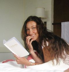 a woman laying in bed reading a book with her hand on her chin and smiling