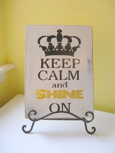 a sign that says keep calm and shine on with a crown in the center, against a yellow wall