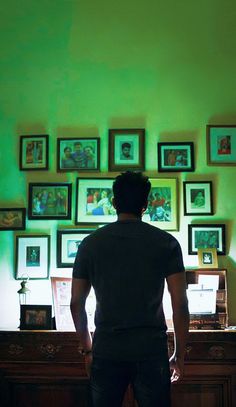 a man standing in front of a green wall with many pictures on the wall behind him