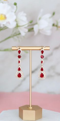 These earrings showcase a pear-shaped design, accentuated with ruby red cubic zirconia (CZ) stones that exude a rich, vibrant hue. They dangle gracefully, adding a pop of color and elegance to any ensemble, making them a perfect statement piece for special occasions or everyday wear. Length: 2 inches Width: 0.50 inches Closure: Bullet with Disc Backs Material: Brass with 18K Gold Plating with Rhodium Coating Lead Free and Hypoallergenic Elegant Red Drop Crystal Earrings, Red Prom Accessories, Red Teardrop Earrings For Formal Occasions, Red Drop Earrings For Formal Occasions, Red Ruby Drop Earrings, Red Dangle Teardrop Earrings For Formal Occasions, Red Teardrop Dangle Earrings For Formal Occasions, Red Teardrop Chandelier Earrings As Gift, Red Pear-shaped Earrings For Formal Occasions