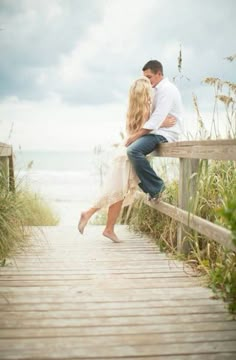 a man and woman are sitting on a wooden bridge by the beach looking at each other