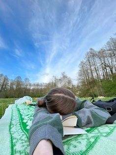 a woman laying on top of a green blanket next to a lush green forest under a blue sky