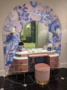 a pink vanity and stool in front of a mirror with blue flowers on the wall