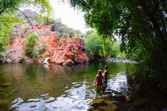 two people are wading in the water near some red rocks and green trees on either side