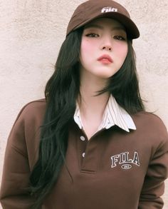 a woman with long black hair wearing a brown shirt and hat, standing in front of a white wall