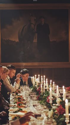 a group of people sitting around a table with candles in front of them and a painting on the wall behind them