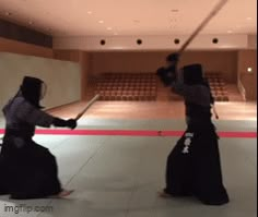 two people in black outfits are playing with swords inside an empty room filled with seats