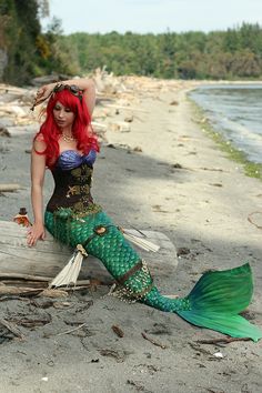 a woman with red hair is sitting on the beach next to a mermaid tail and body