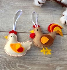 two felt rooster ornaments hanging from a twig on a wooden table next to cotton balls