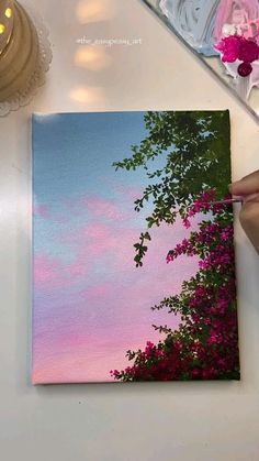 someone is painting the sky and trees with pink flowers in front of them on a white table