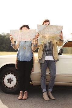 two people standing next to a car holding up maps