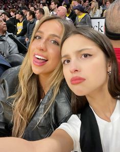 two women taking a selfie in front of an audience at a basketball game, one with her arm around the other's shoulder