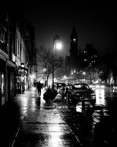 a city street at night with cars parked on the side and people walking down the sidewalk