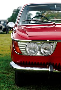 the front end of an old red car parked on some grass in a field with other cars behind it