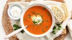 a bowl of tomato soup with bread and sour cream on a woven place mat next to it