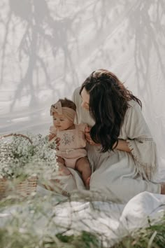 a woman holding a baby in her arms while sitting on the ground next to flowers