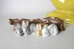 three small white rabbits sitting next to each other on top of a wooden branch in front of a yellow vase