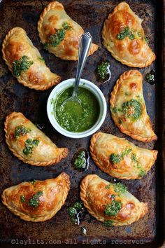 several pastries on a baking sheet with a bowl of green sauce and spoon in the middle