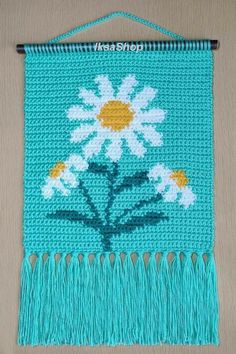 a crocheted wall hanging with white daisies on blue yarn and fringes