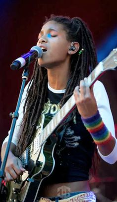 a woman with dreadlocks singing into a microphone and holding a guitar in front of her