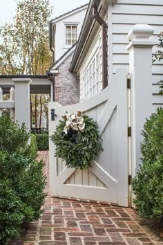 a white gate with a wreath hanging on it's side and brick walkway leading to the front door