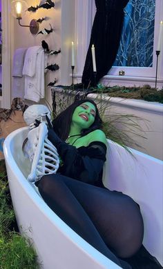 a woman with green makeup sitting in a bathtub next to a halloween decoration and candles