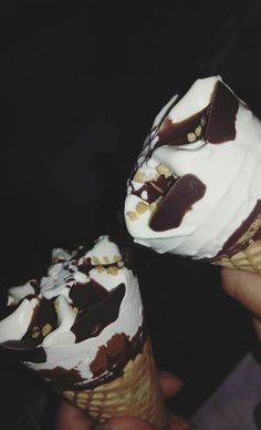 two ice cream cones with chocolate and nuts on top, being held by someone's hand