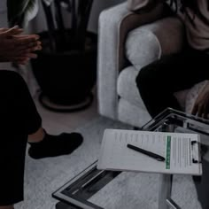 two people sitting on couches in a living room, one holding a pen and the other writing