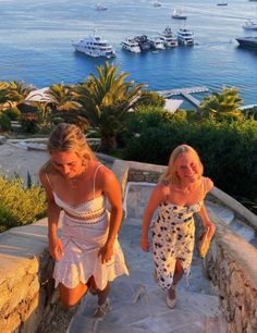 two women are walking up some steps near the ocean and boats in the water behind them