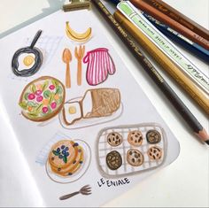 some colored pencils are laying on top of a drawing book with food and utensils