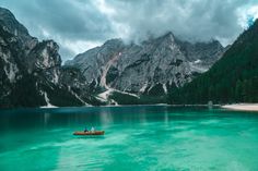 two people in a small boat on a lake surrounded by mountains and pine trees, under cloudy skies