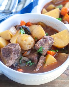 two bowls filled with stew and potatoes on top of a wooden table next to silverware