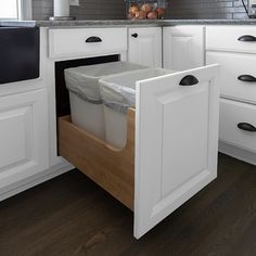 a kitchen with white cabinets and drawers has two trash cans in the bottom half of the cabinet